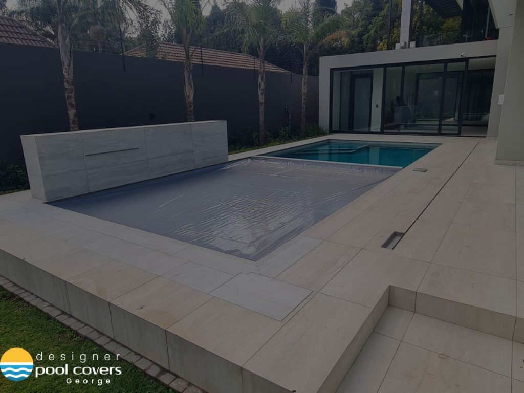 designer pool covers A modern backyard featuring a sleek swimming pool, partially shielded by an elegant retractable pool cover, is surrounded by a tiled deck and swaying palm trees, ensuring both style and pool protection.